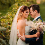 A bride in a strapless white wedding gown with a long veil and a bouquet of pale pink and cream flowers, hugging a groom in a navy suit in a vineyard at golden hour.