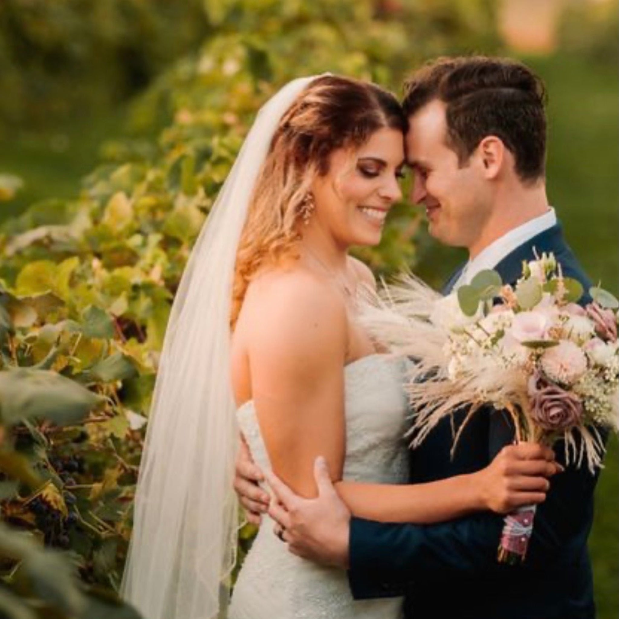 A bride in a strapless white wedding gown with a long veil and a bouquet of pale pink and cream flowers, hugging a groom in a navy suit in a vineyard at golden hour.