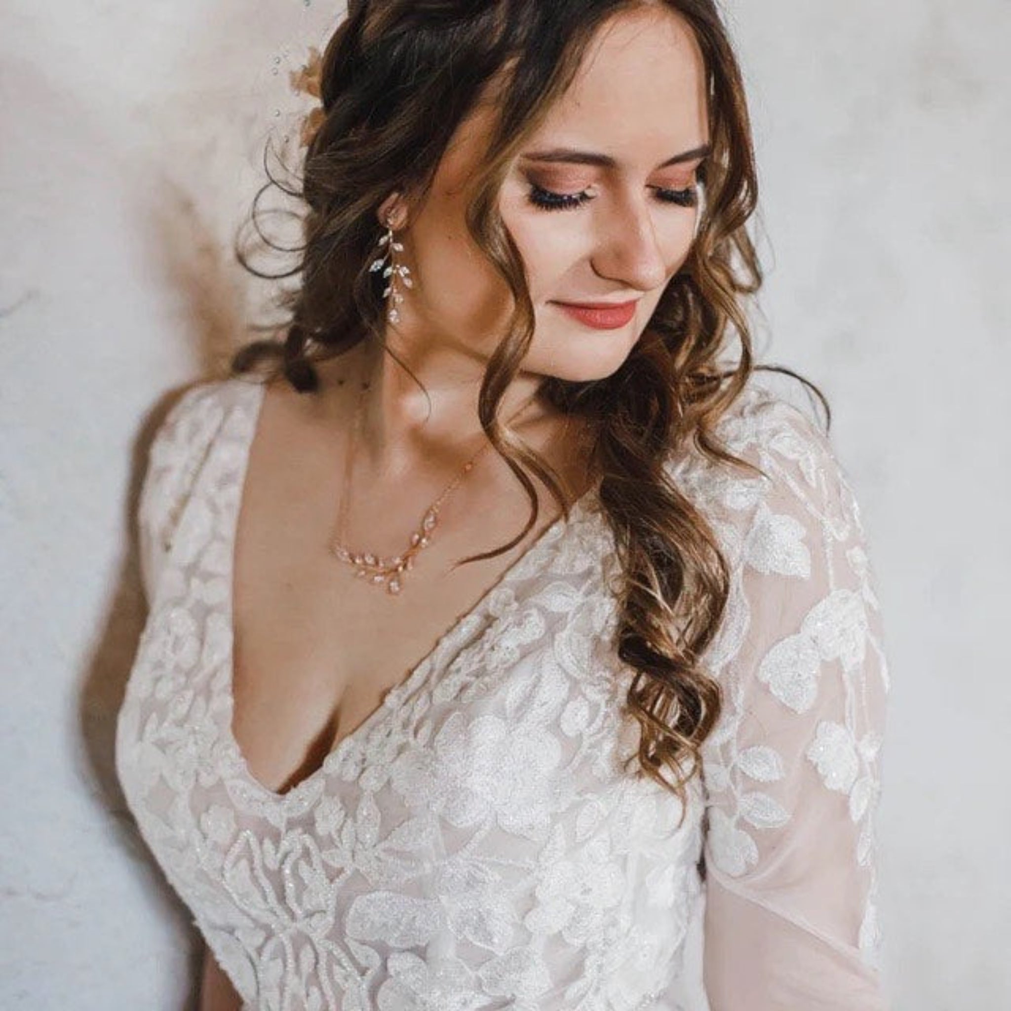 A woman wearing a white lace wedding dress with floral embroidery and sheer long sleeves, styled with a delicate rose gold necklace and matching earrings against a soft, textured off-white background.