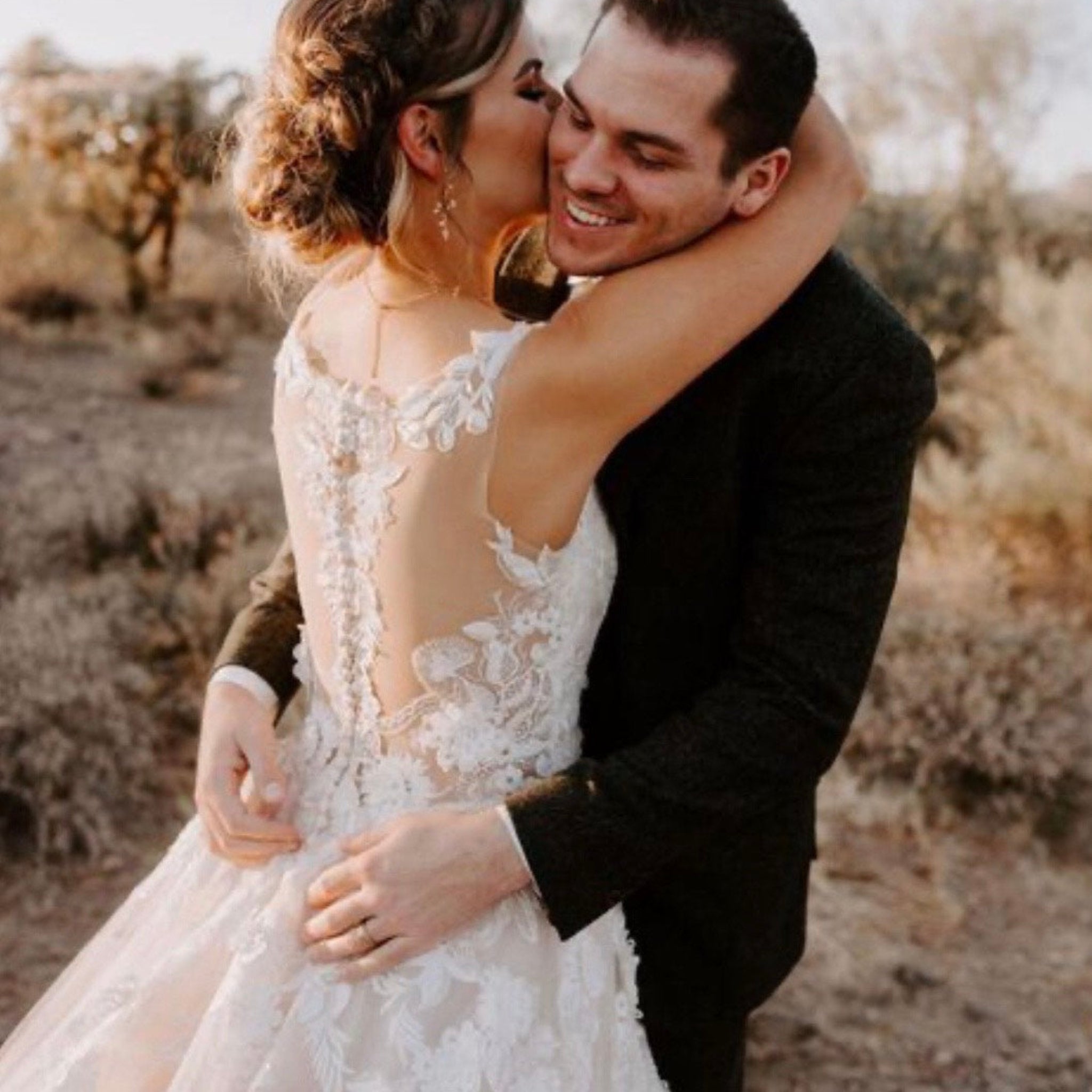 A smiling bride in a white lace wedding dress with an open back, embraced by a groom in a black suit outdoors in a dry, sunlit landscape.