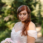 A woman outdoors wearing a white off-the-shoulder lace dress, a floral headpiece, and dangling earrings, holding a bouquet of pale pink flowers against a blurred green garden background.