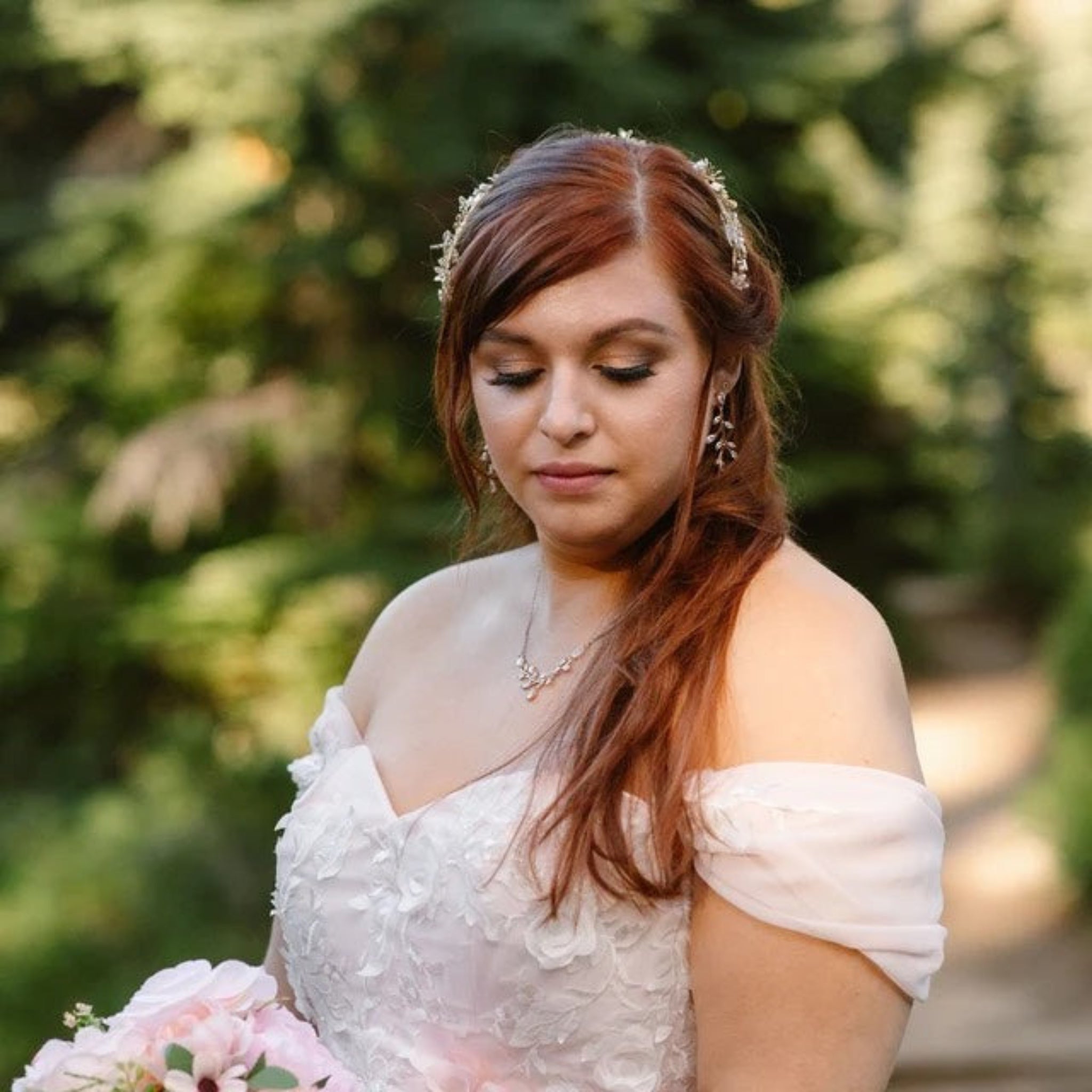 A woman outdoors wearing a white off-the-shoulder lace dress, a floral headpiece, and dangling earrings, holding a bouquet of pale pink flowers against a blurred green garden background.