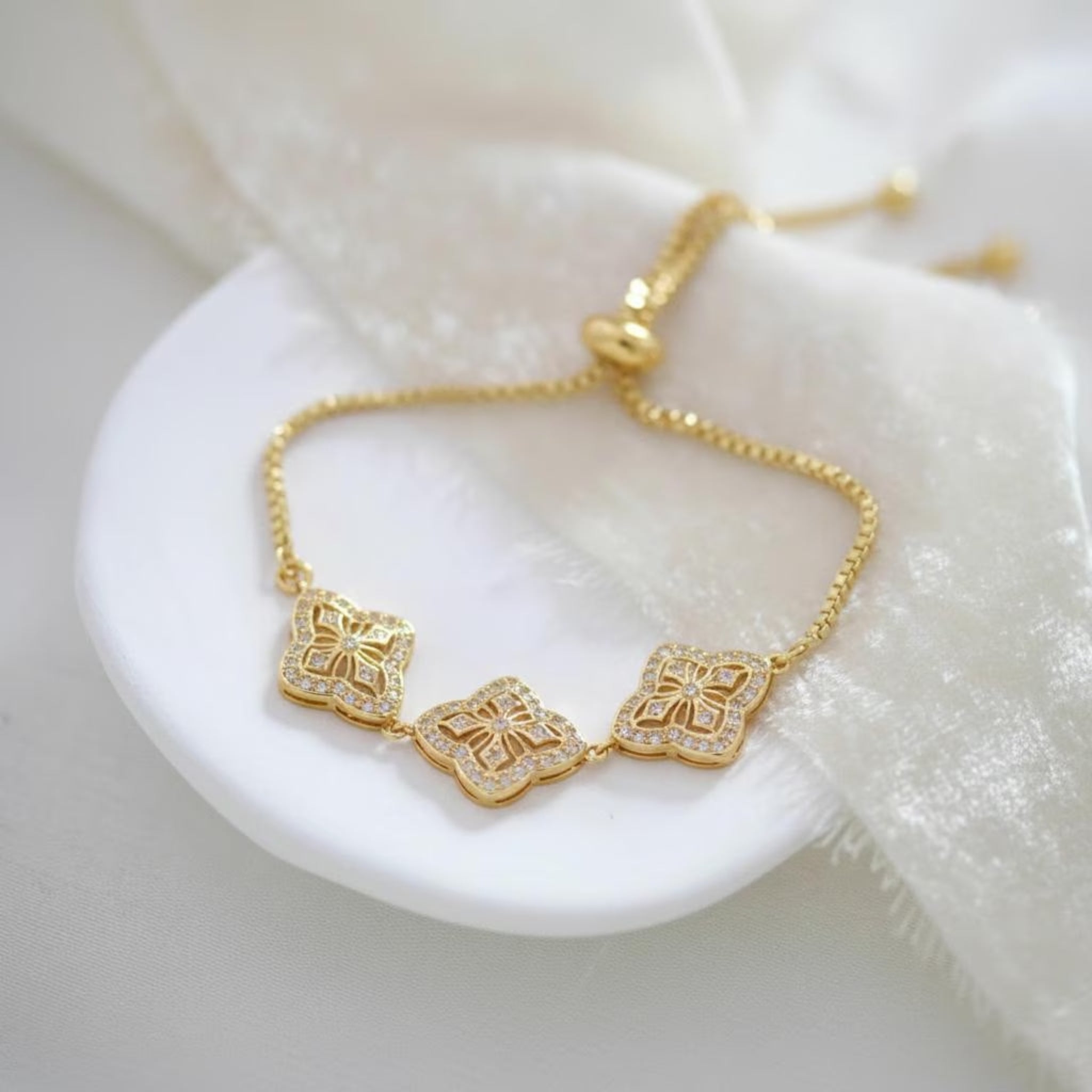 Gold Addia bridal bracelet featuring sparkling cubic zirconia quatrefoil motifs and an adjustable slider chain, elegantly displayed on a white ceramic dish.