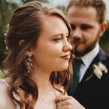 A bride wearing delicate silver leaf drop earrings and a ring, looking to the side with a groom in a dark suit and green tie softly blurred in the background.