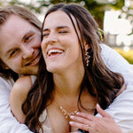 A smiling couple embracing outdoors, with the woman wearing a delicate gold leaf necklace and matching earrings, and the man wearing a white shirt.