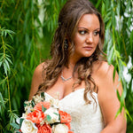 A woman in a strapless white lace wedding gown holding a bouquet of coral, white, and green flowers, framed by hanging green leaves.
