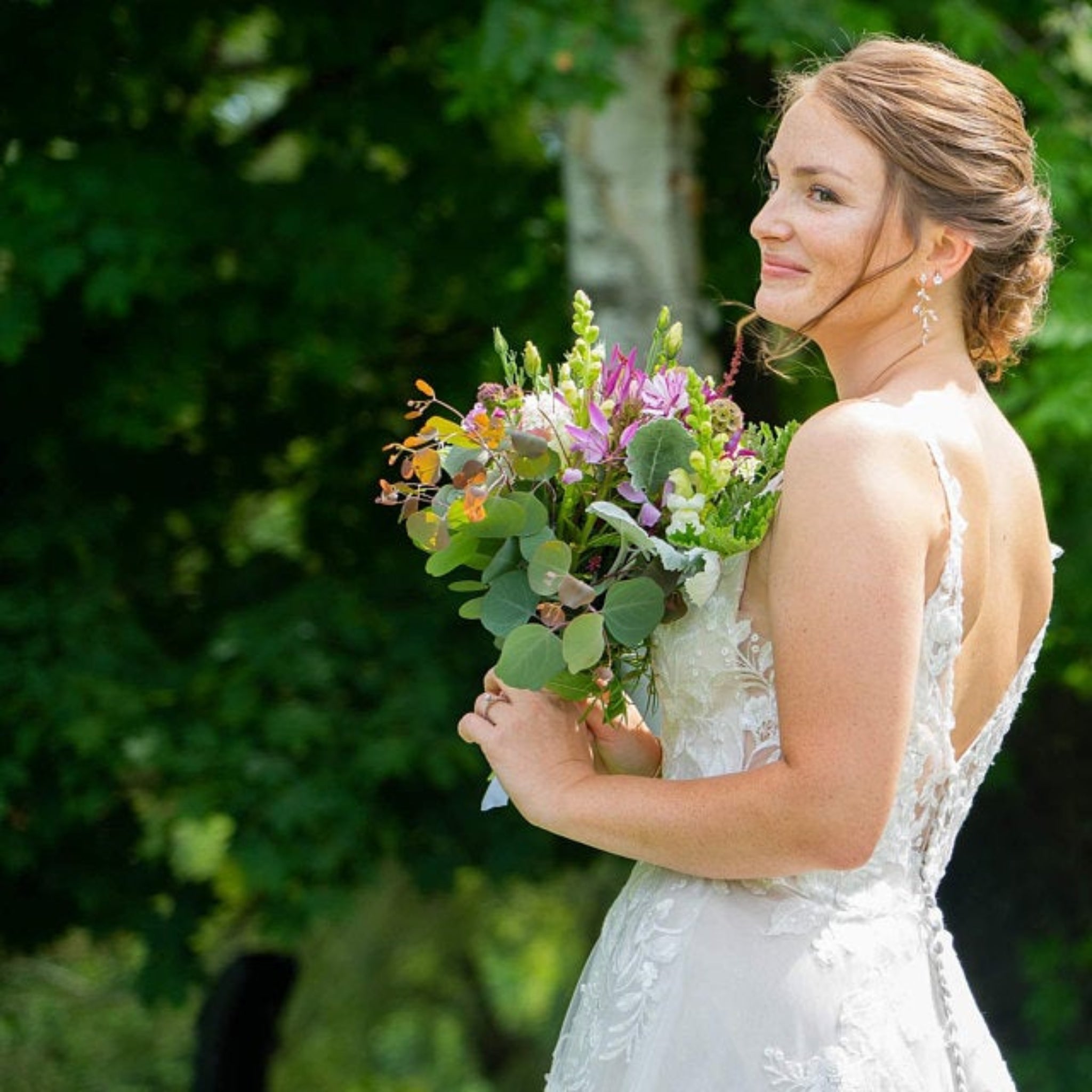 A bride in a white lace wedding dress holding a colorful wildflower bouquet, photographed outdoors against a blurred green forest background.