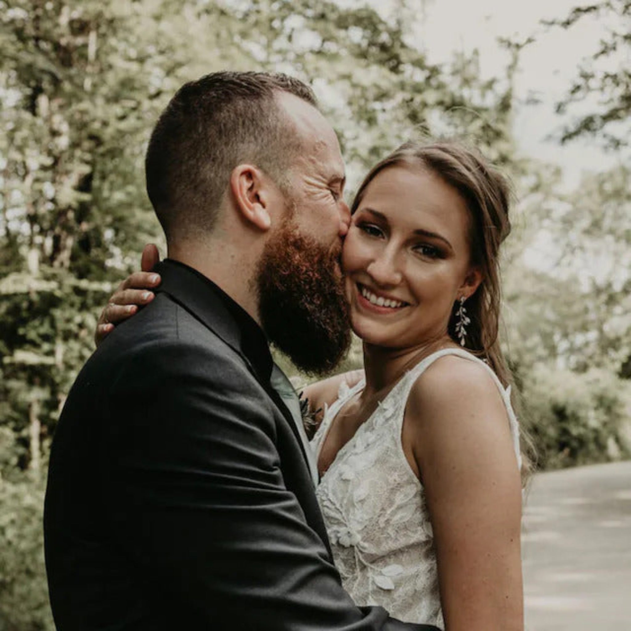A smiling bride in a white lace sleeveless wedding dress and a groom in a black suit kiss on the cheek outdoors with trees in the background.