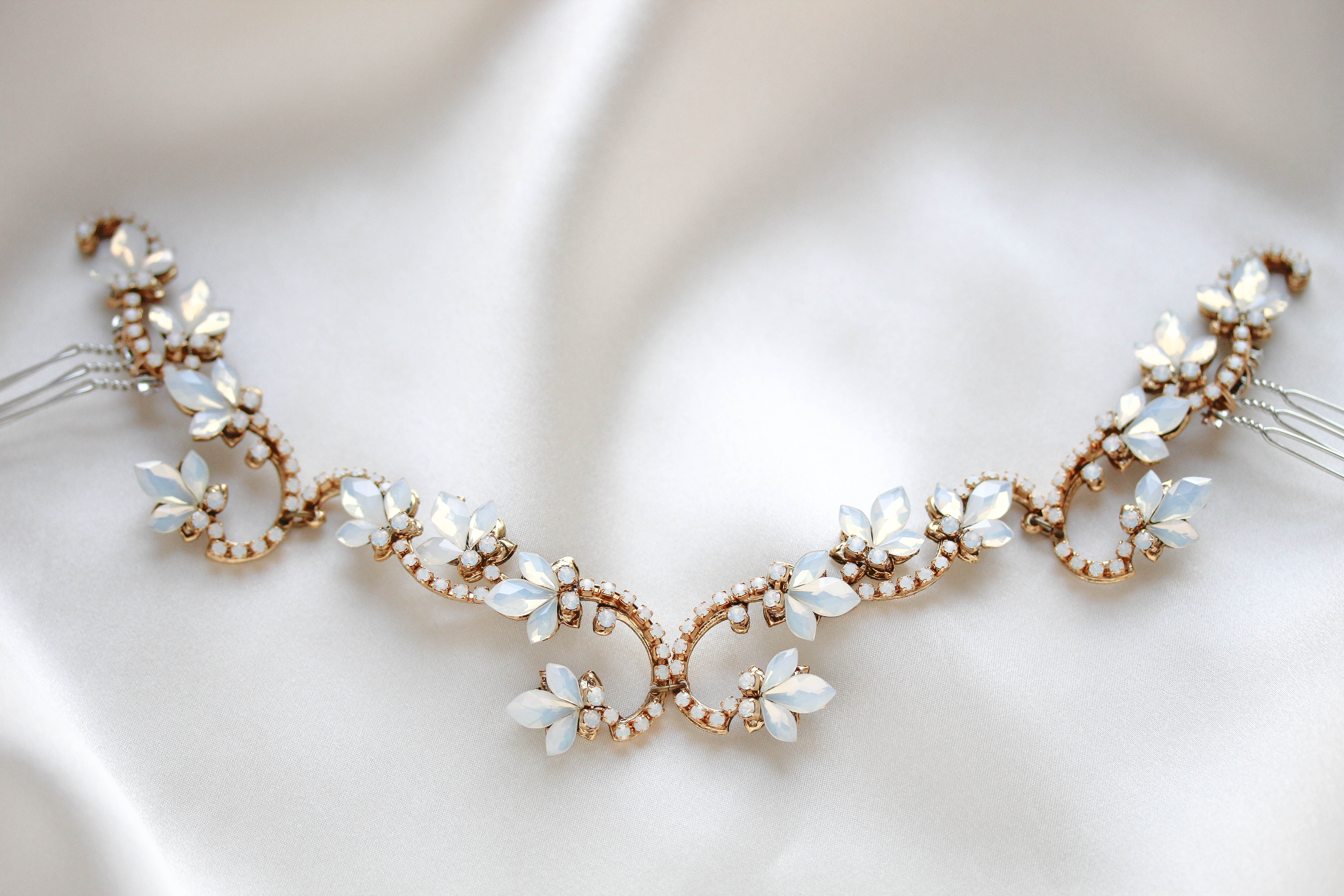 Close-up of Cleo antique gold bridal hair vine featuring Swarovski white opal crystal flowers, intricate sparkling scrollwork and hair comb attachments, displayed on soft ivory satin.